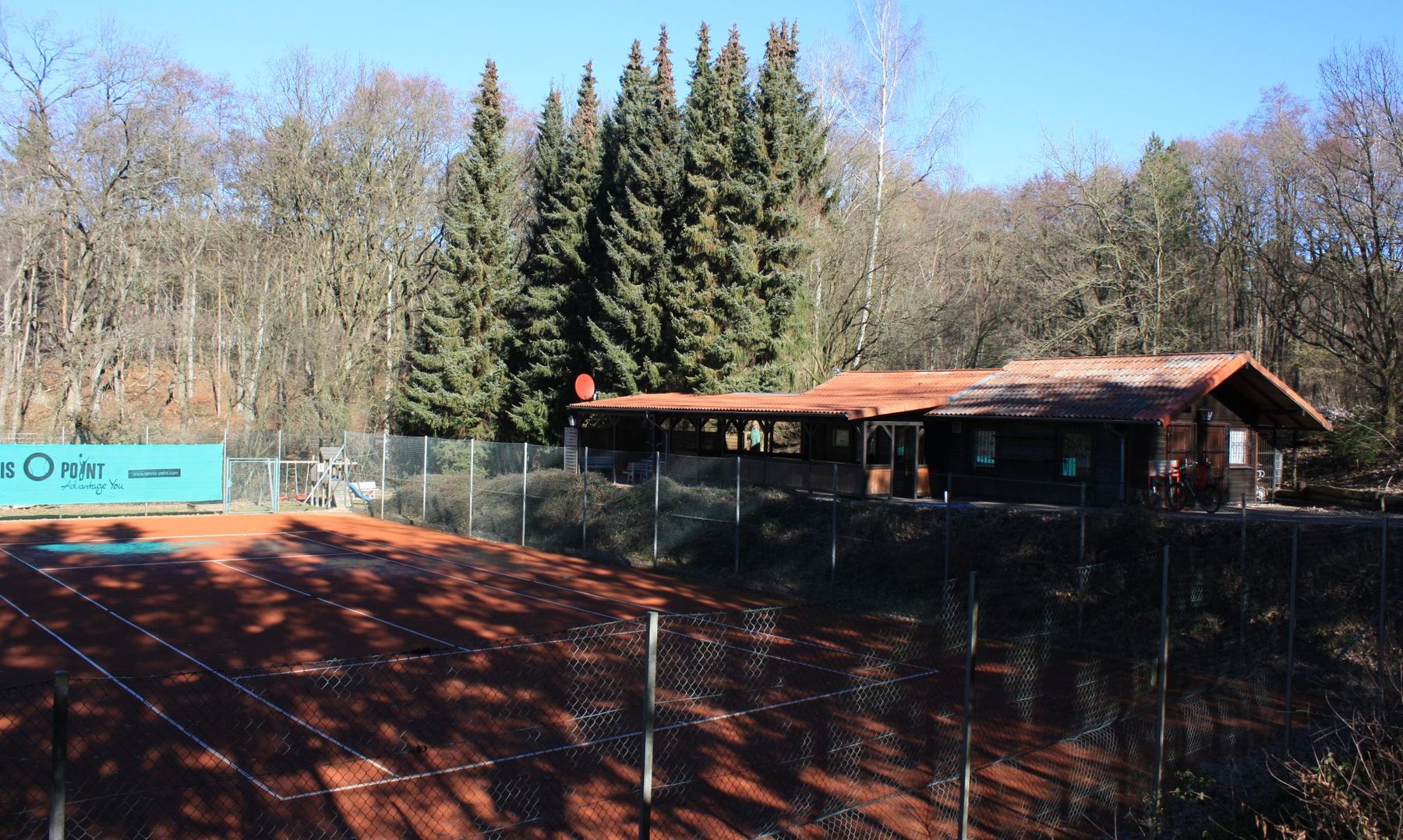 Blick über einen der roten Sandplätze auf das hölzerne Vereinsheim des TC Söhrewald, dahinter Fichten und der Söhrewald.